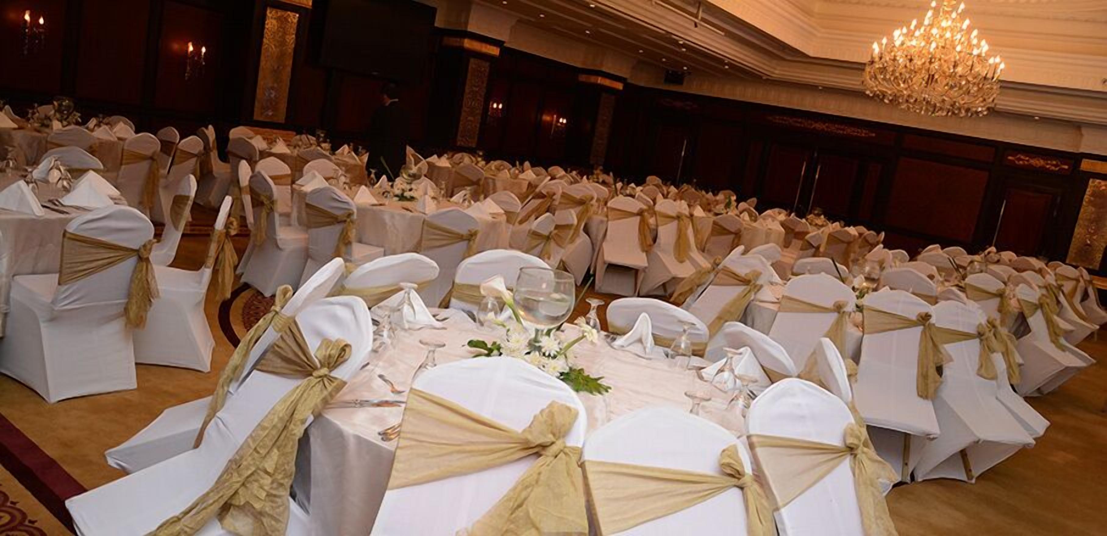 Un salón de banquetes con filas de mesas redondas cubiertas con manteles blancos y fajos dorados, y una araña de cristal colgando del techo.