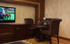 A room with a television displaying a sports event, a wooden cabinet, two leather chairs, and a small table with a vase of flowers.