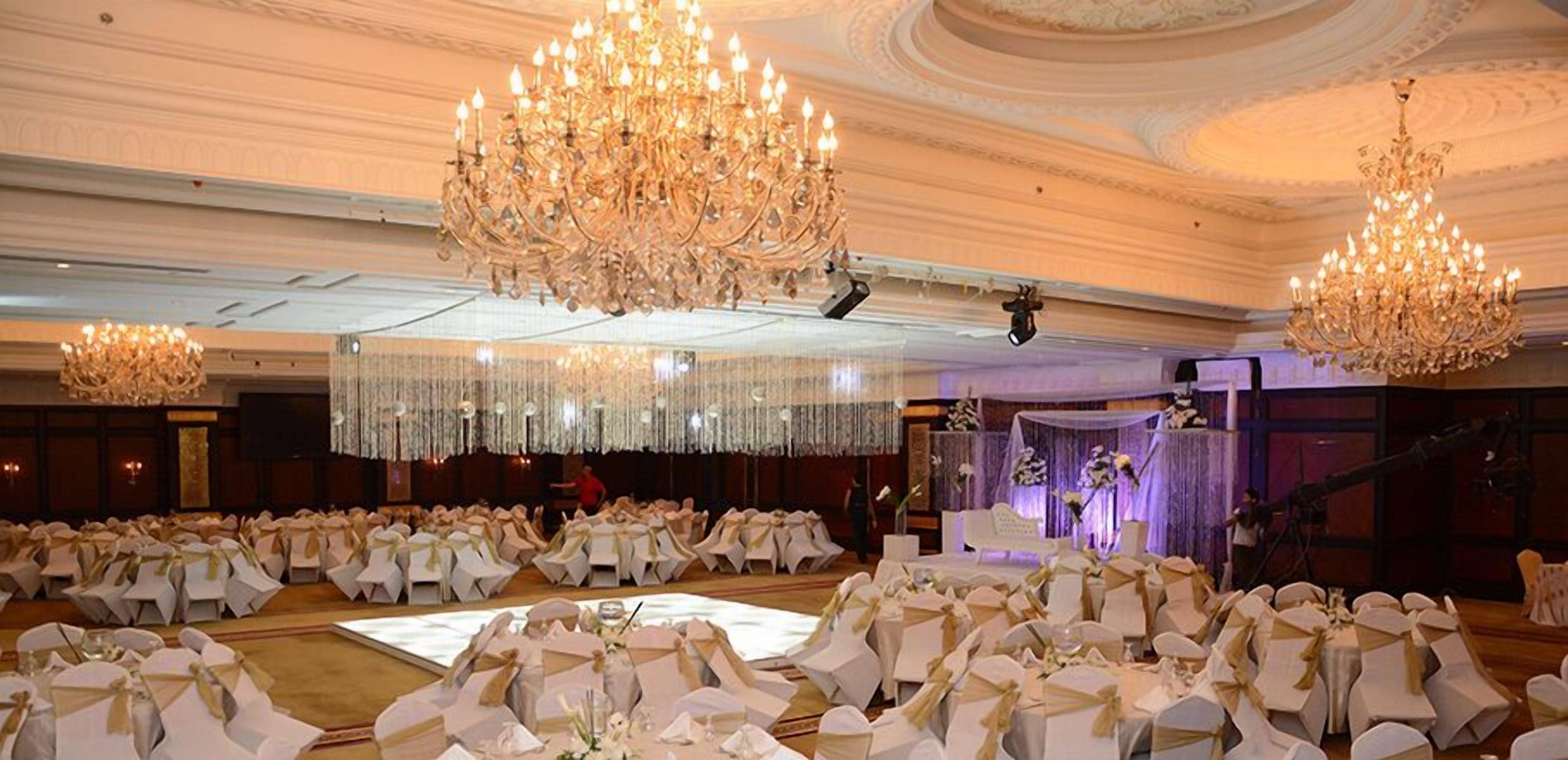 Un salón de banquetes con filas de mesas redondas, grandes candelabros y un escenario con arreglos florales.