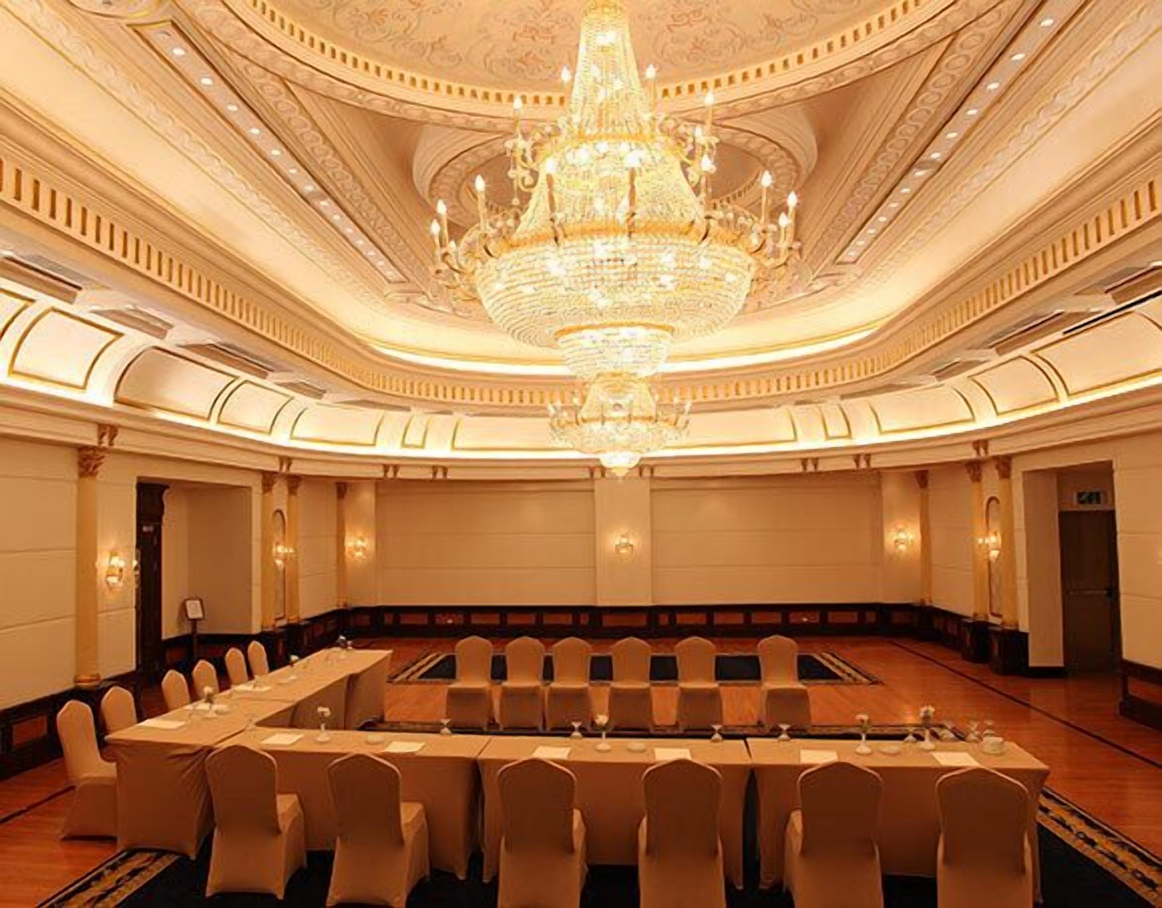 A grand ballroom with a large chandelier, a long conference table, and rows of chairs.