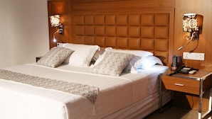 A neatly made bed with patterned pillows, a bedside table with a phone and a lamp, and a wall-mounted light fixture. - Mint Hotel (Lomé)