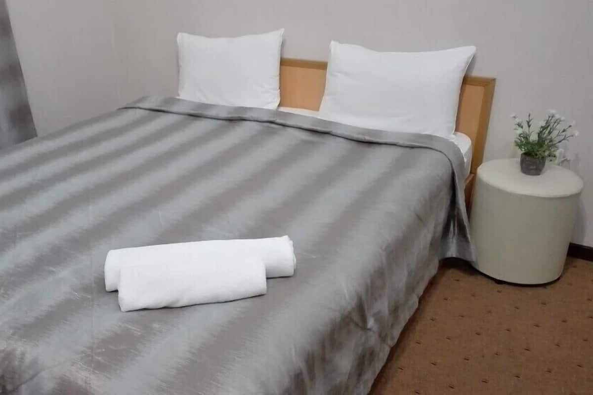 Ein ordentlich bezogenes Bett mit einem gestreiften Überwurf und zwei Kissen, ein runder Nachttisch mit einer Pflanze und ein zur Seite gezogener Vorhang.