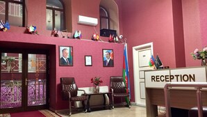 Un área de recepción con mostrador, dos sillones de cuero, una mesita con un jarrón de flores y varias banderas en la pared.