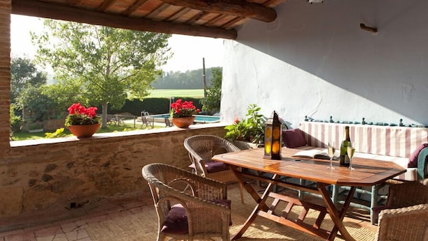 Een terras buiten met rieten meubels, een houten tafel en planten in potten.