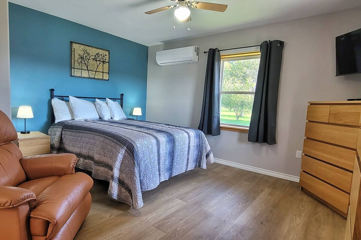 Un dormitorio con cama, una silla, cómoda, ventilador de techo, ventana con cortinas y televisor montado en la pared.