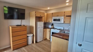 Cocina compacta con gabinetes de madera, microondas, refrigerador, estufa y televisor montado en la pared.