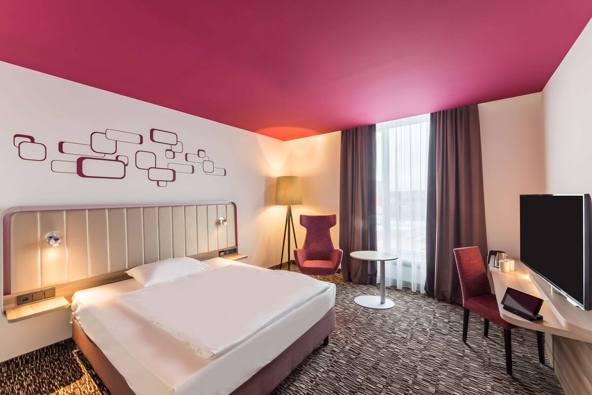 A hotel room with a large bed, a desk with a laptop, a TV, and a wall-mounted lamp.