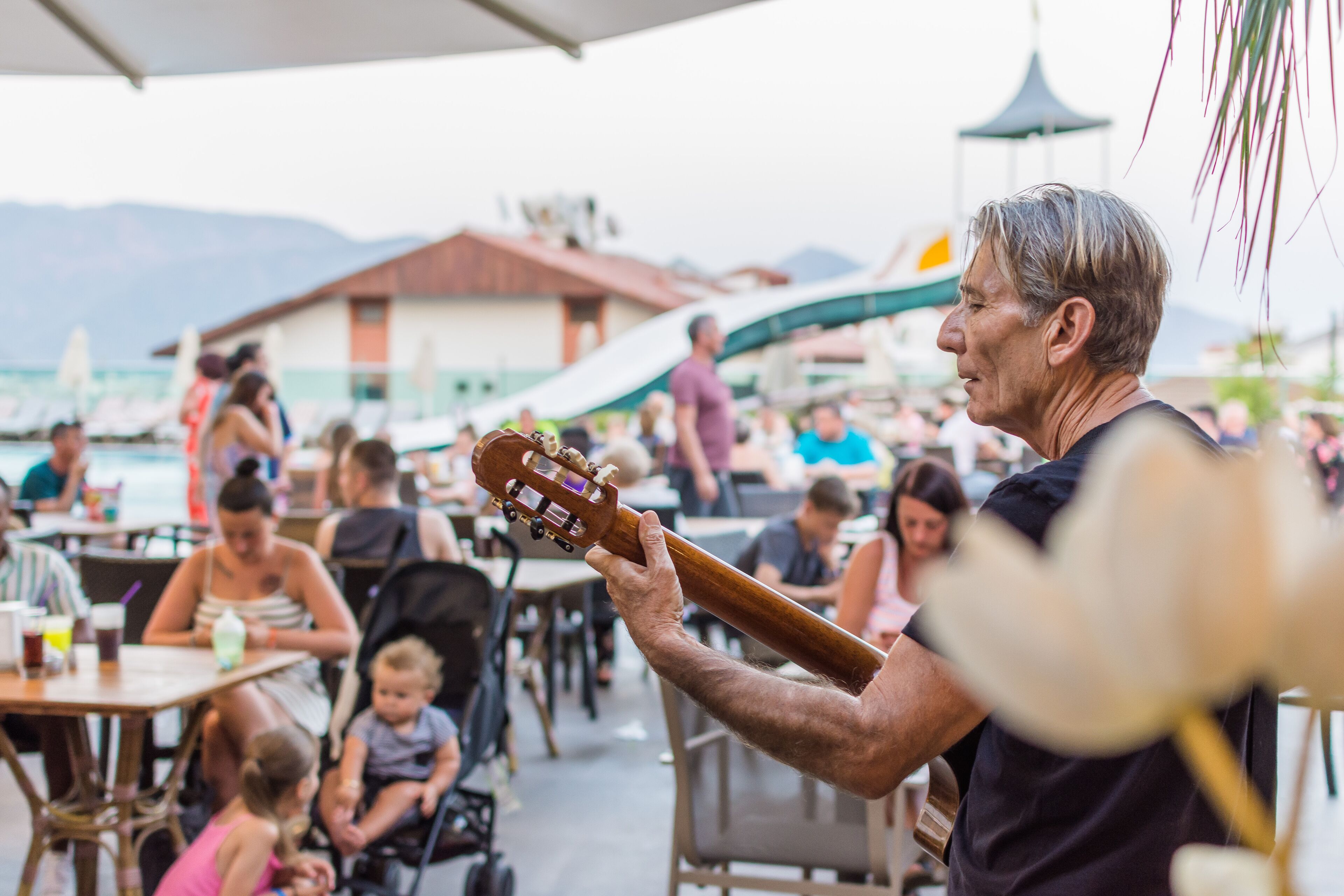 Een man speelt gitaar tijdens een evenement buiten, met tafels en stoelen, een gebouw op de achtergrond en bergen in de verte.