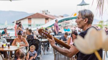 Een man speelt gitaar tijdens een evenement buiten, met tafels en stoelen, een gebouw op de achtergrond en bergen in de verte.