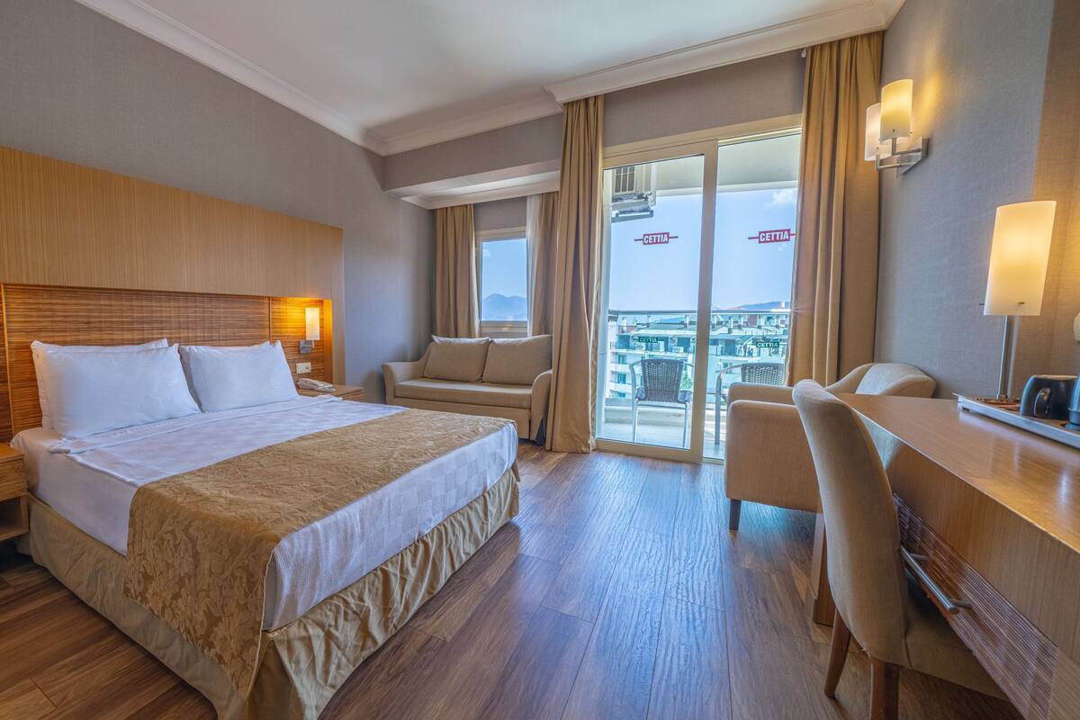 A hotel room with a large bed, a desk, a chair, a sofa, and a balcony with a view.