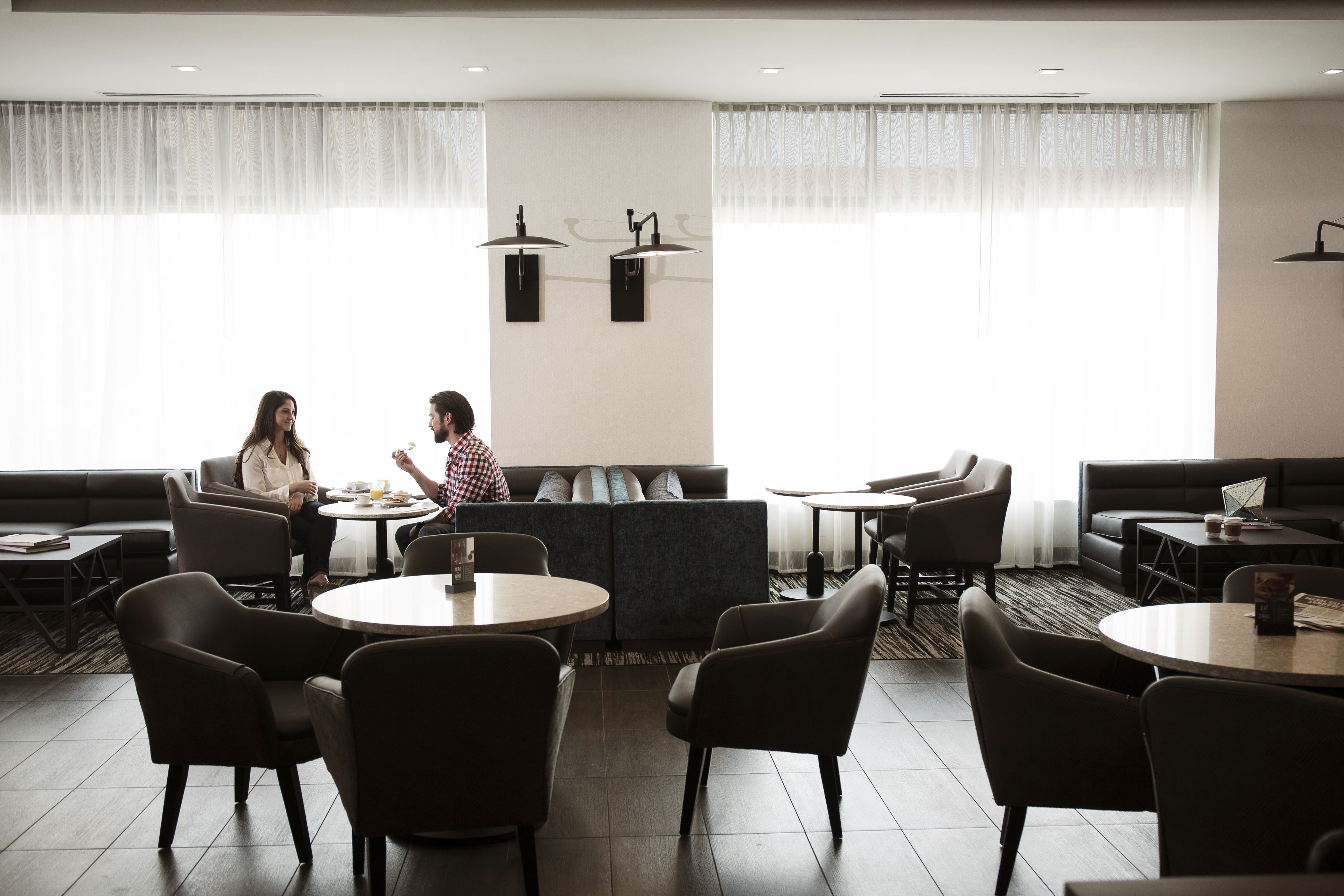 A modern hotel lobby with round tables, comfortable seating, and large windows with sheer curtains.