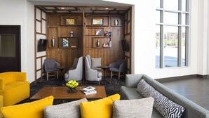 A modern lobby with a wooden panel wall, seating area with sofas and armchairs, a coffee table with books and fruit, and large windows offering a view. - Hyatt Place Lubbock (Lubbock)