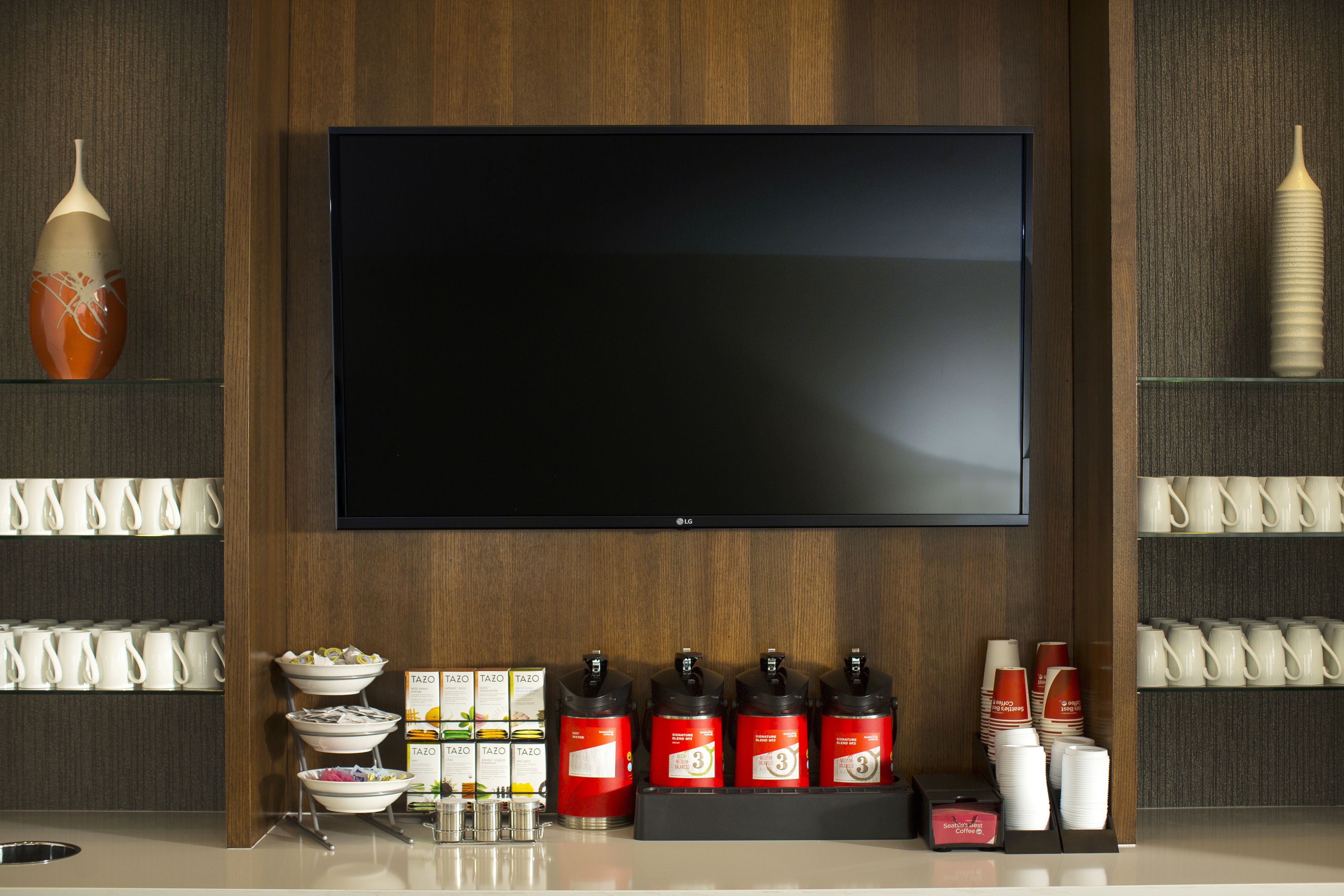 Una habitación de hotel moderna con un televisor de pantalla plana grande montado en la pared, una zona de café con varias tazas y una zona de té con bolsitas y tazas.