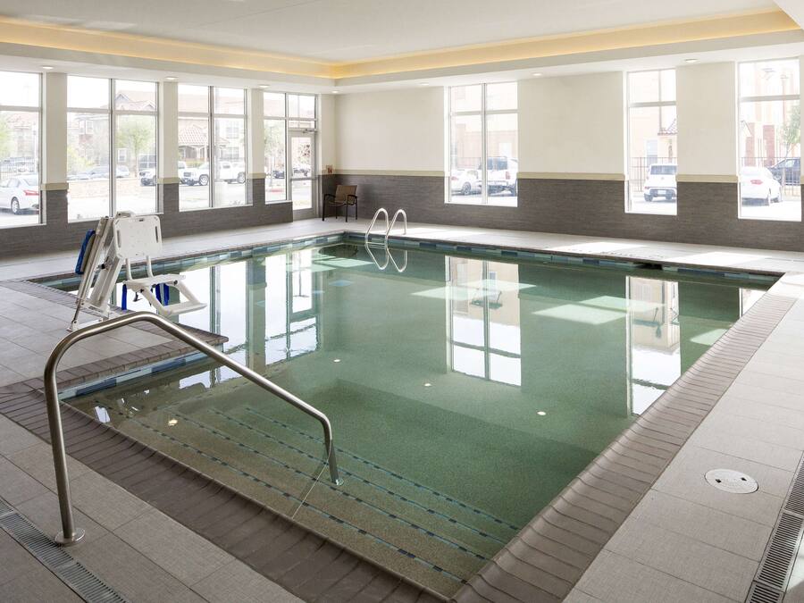 An indoor swimming pool with clear glass walls, a metal pool ladder, and a view of parked cars outside.
