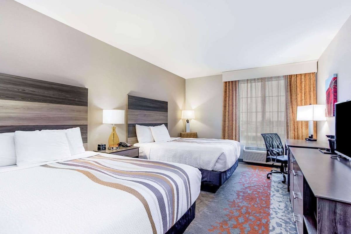 Ein Hotelzimmer mit zwei Betten, einem Schreibtisch, einem Stuhl, einem Fernseher und einem großen Fenster mit Vorhängen.