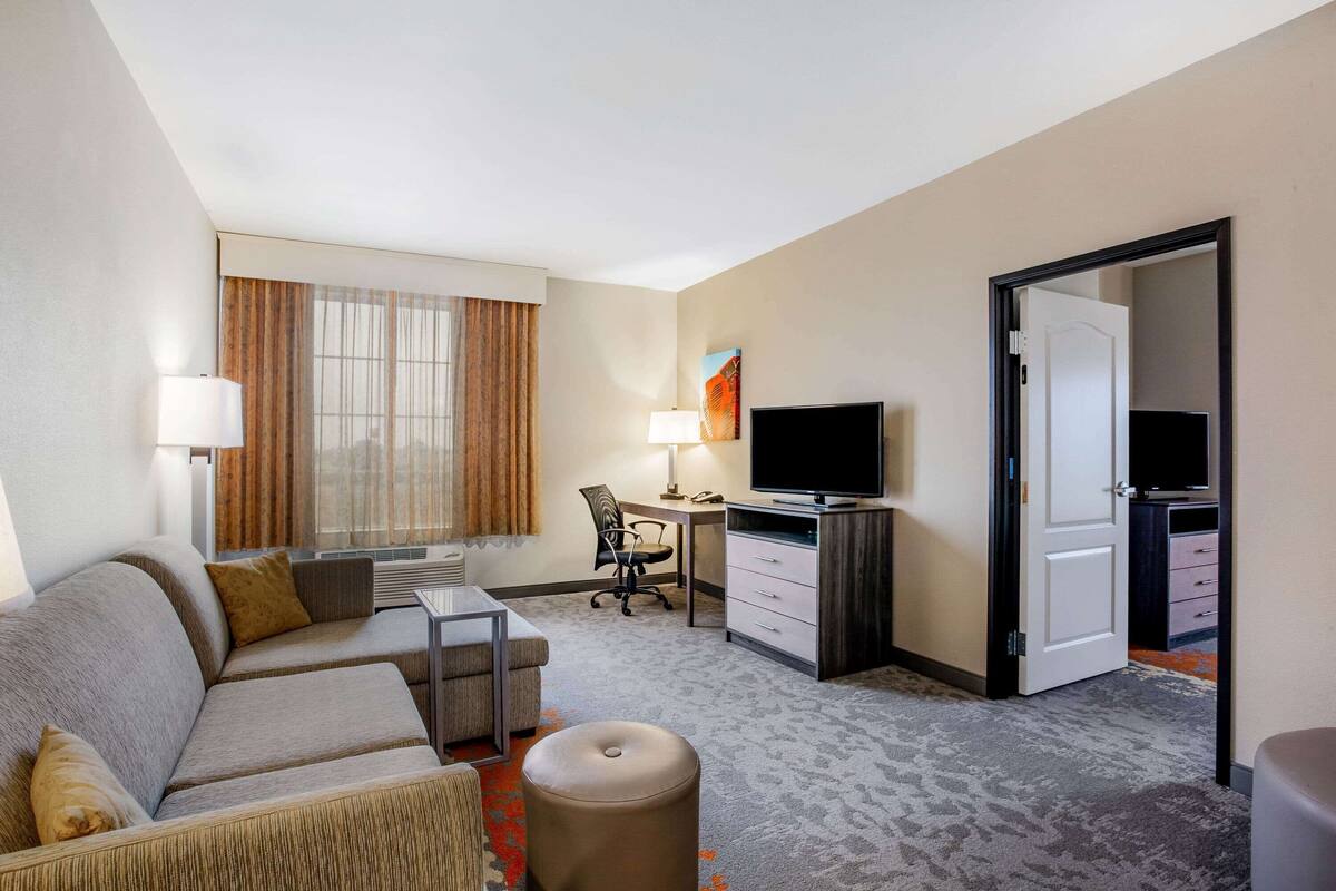 Ein Hotelzimmer mit einer Couch, einem Schreibtisch mit Computer, einem Fernseher und einer Tür zu einem weiteren Raum.