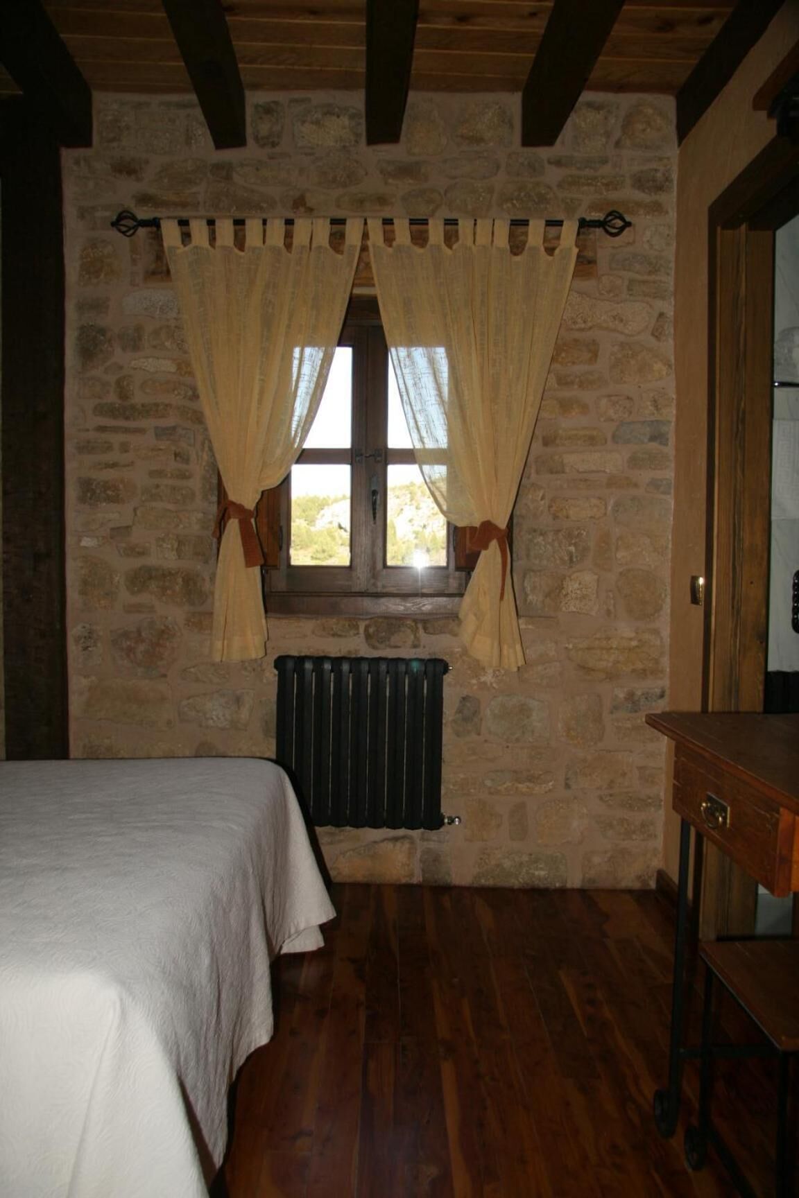 A bedroom with a stone wall, a window with curtains, a radiator, a bed, and a wooden table.