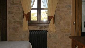 A bedroom with a stone wall, a window with curtains, a radiator, a bed, and a wooden table.