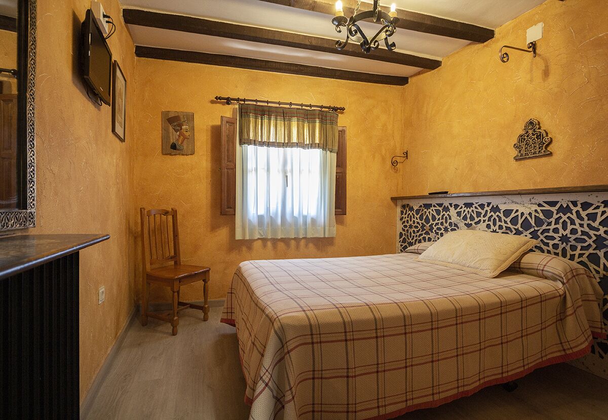 A bedroom with a bed, a chair, a window with curtains, a wall-mounted TV, and a decorative headboard.