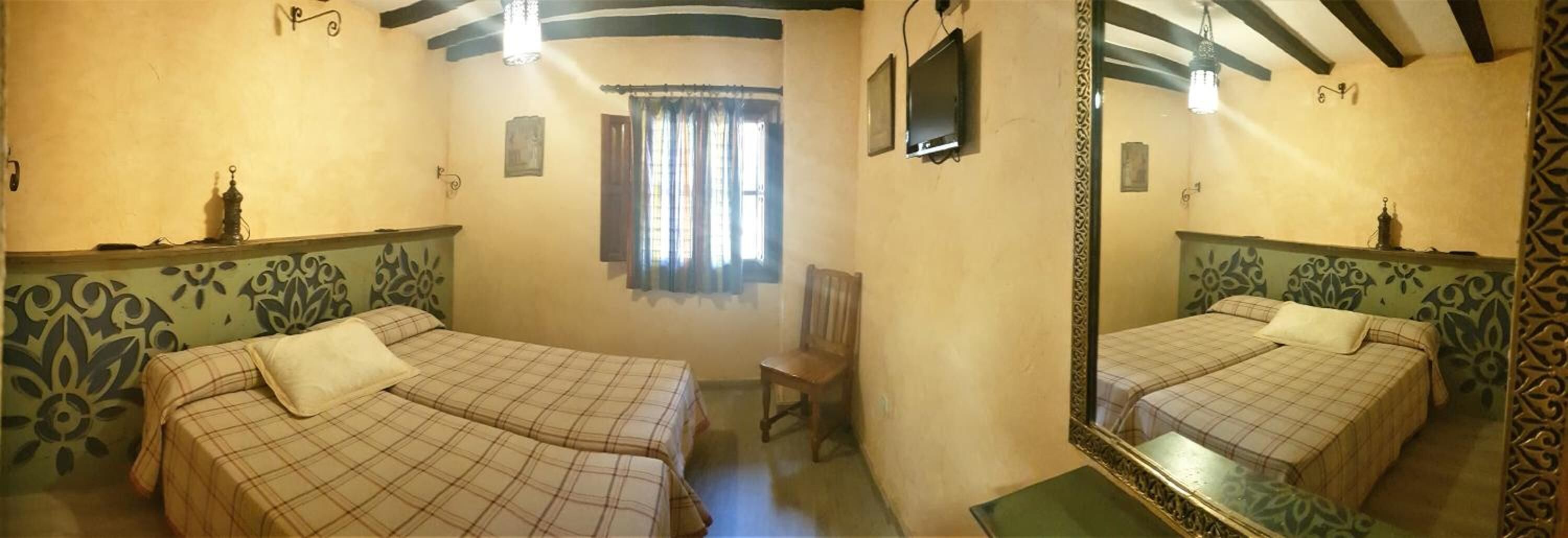 Una habitación con dos camas, una silla, un ventanal con cortinas, un televisor de pantalla plana y un espejo.