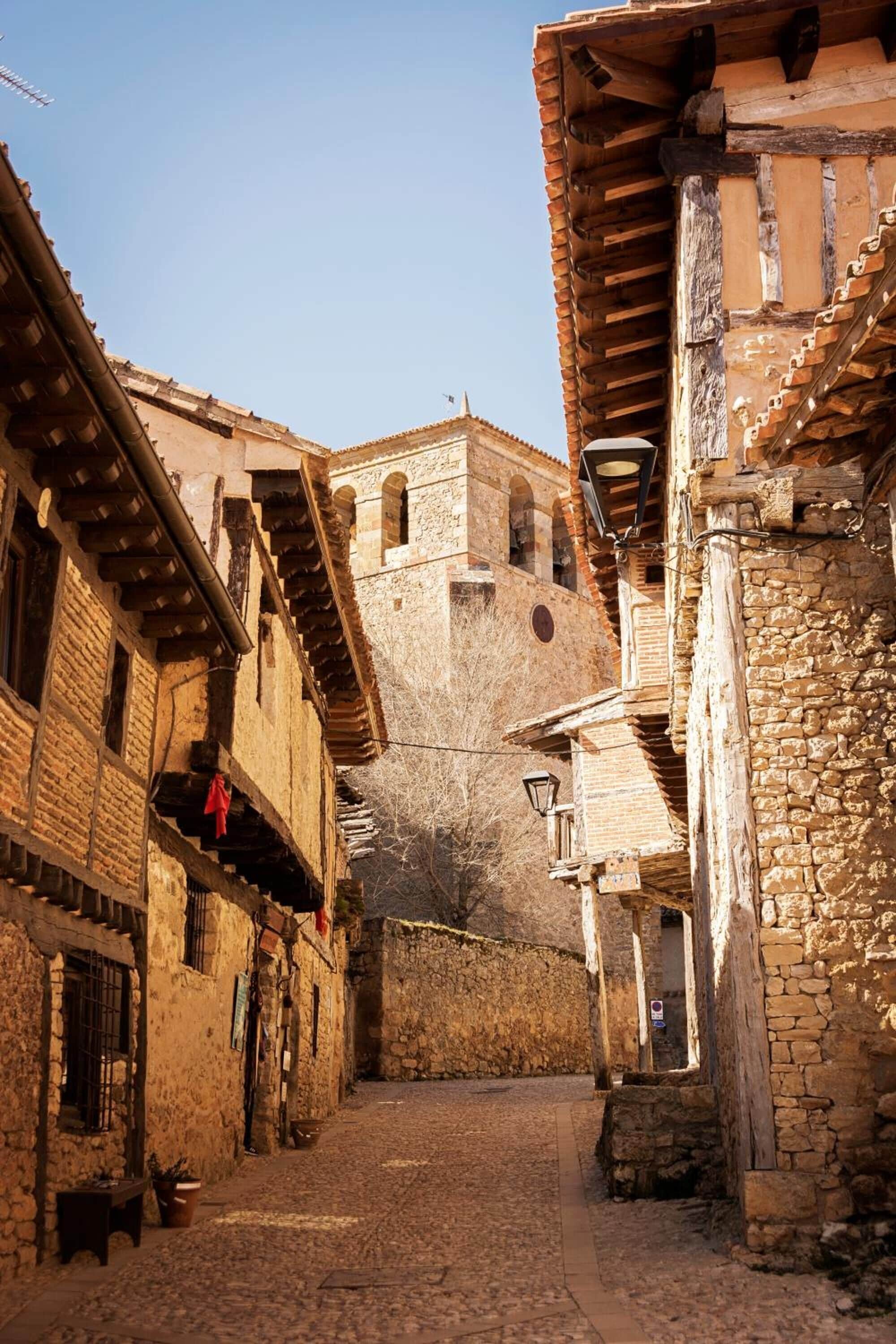 A narrow cobblestone street flanked by historic stone buildings with wooden balconies and a prominent tower in the background.