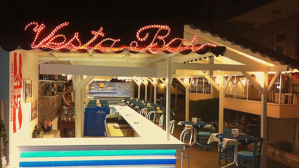Um bar à beira-mar com um letreiro de neon, mesas ao ar livre e um balcão visível.