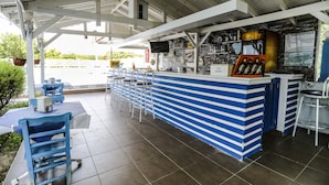 Uma área de bar com balcão azul e branco listrado, vários bancos e piso ladrilhado.