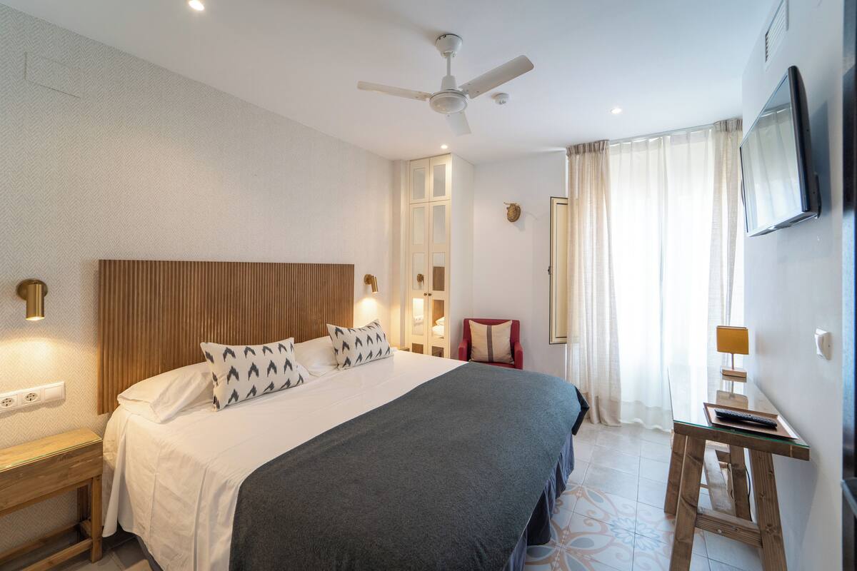 A hotel room with a large bed, a ceiling fan, a TV mounted on the wall, and a window with curtains.