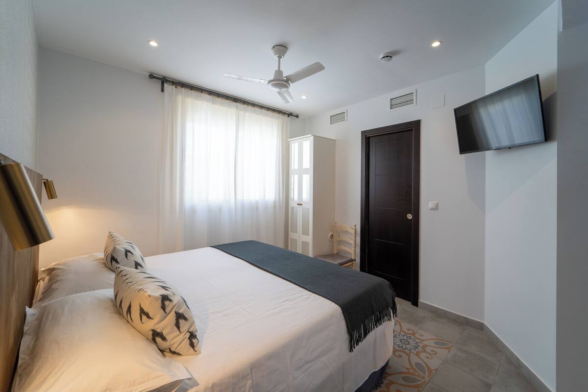 A hotel room with a bed, a ceiling fan, a TV mounted on the wall, and a door.