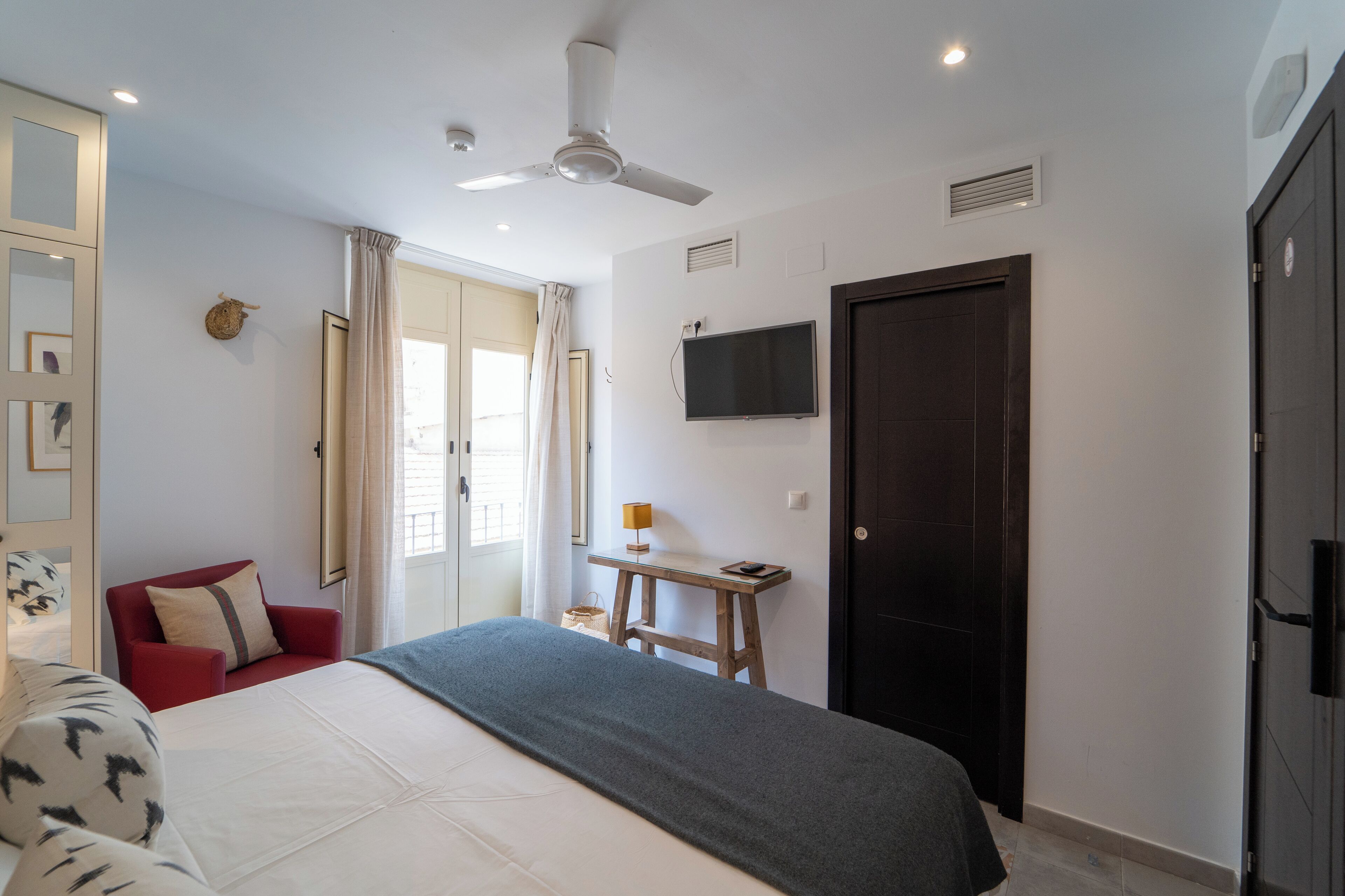 Una habitación de hotel con una cama, un sillón rojo, una mesita de madera con una lámpara, un televisor montado en la pared y un ventilador de techo.