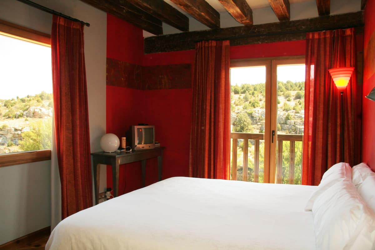 A bedroom with a bed, a television on a small table, and a balcony with a view of greenery.