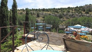 A patio area with stone flooring, metal chairs, and tables. There is a view of a hilly landscape with trees and a building in the background.