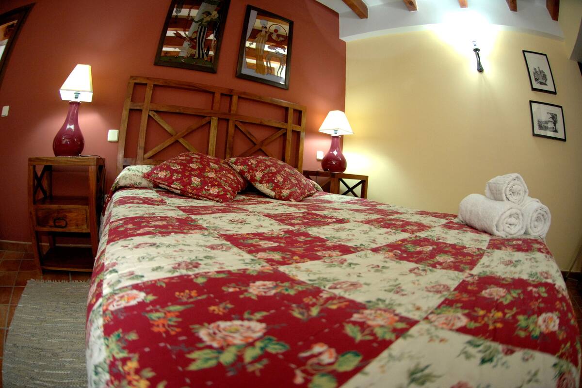 Ein Bett mit floraler Steppdecke, zwei Nachttischlampen, einem Holz-Kopfteil und einem zusammengerollten Handtuch daneben.