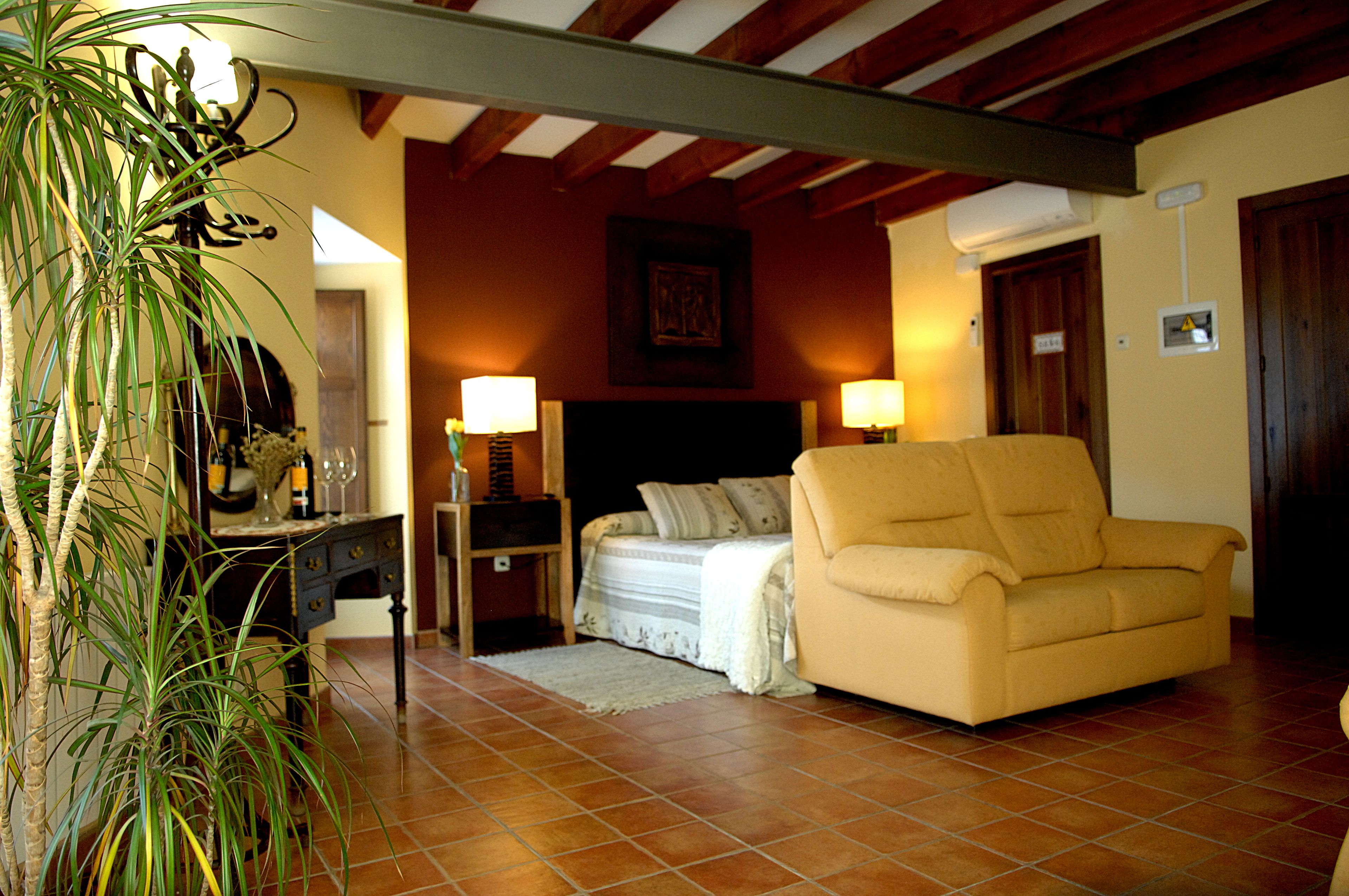 A living room with a sofa, a bed with a headboard, a nightstand with a lamp, a potted plant, and a tiled floor.