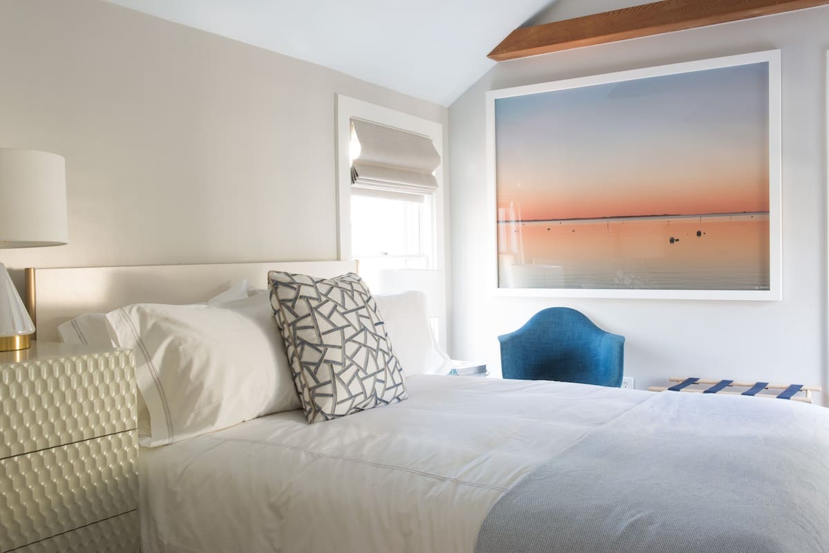 Ein Hotelzimmer mit Bett, Nachttisch, Lampe, Fenster mit Sonnenuntergangsblick und einem blauen Sessel.