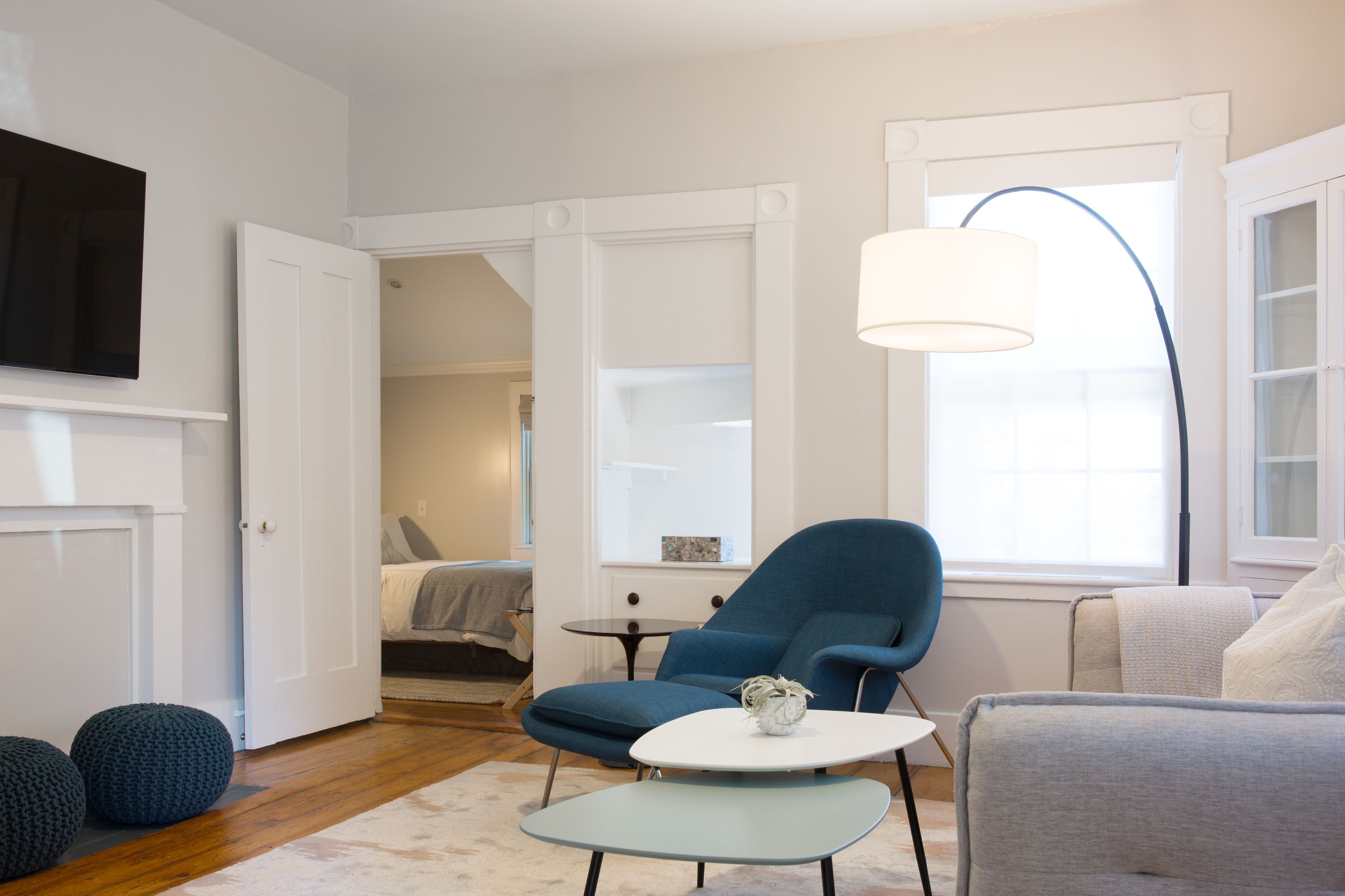 Een moderne woonkamer met een blauwe fauteuil, een witte salontafel en een grijze bank. Er is een televisie aan de muur bevestigd, een raam met een lamp, en een deur die naar een andere kamer leidt.