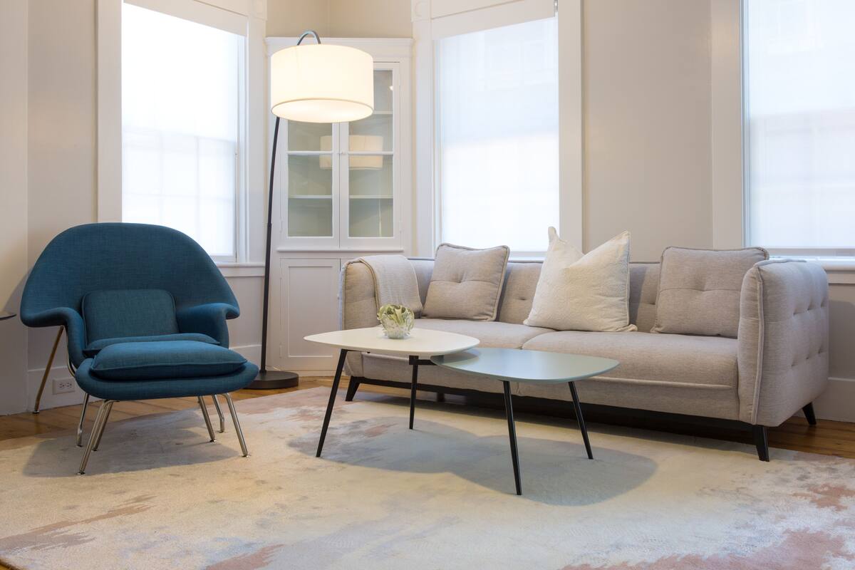 Ein Wohnzimmer mit blauem Sessel, beiger Couch und kleinem weißen Tisch.