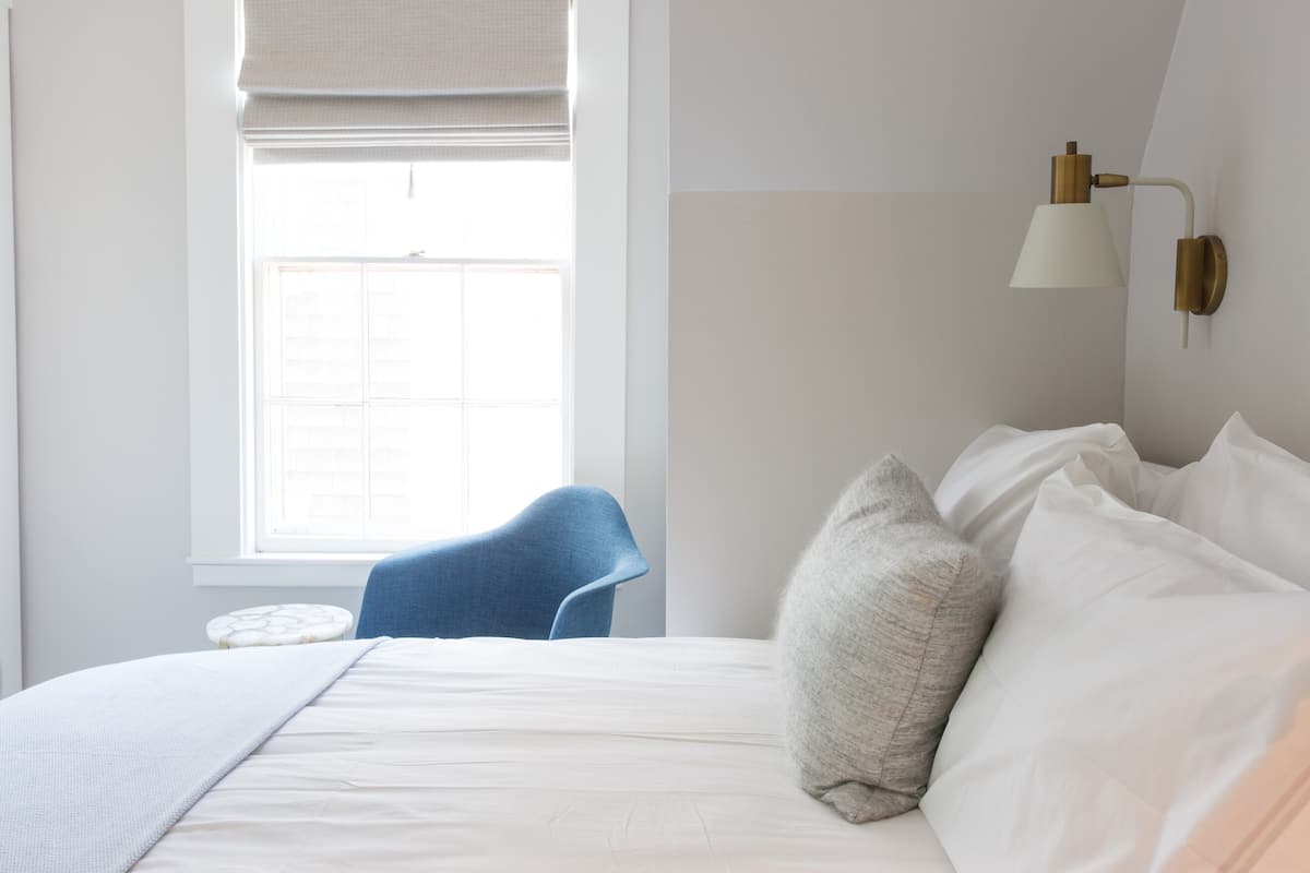 Ein Schlafzimmer mit Bett, Kissen, einem blauen Sessel, einem kleinen runden Tisch und einem Fenster mit weißem Rollo.