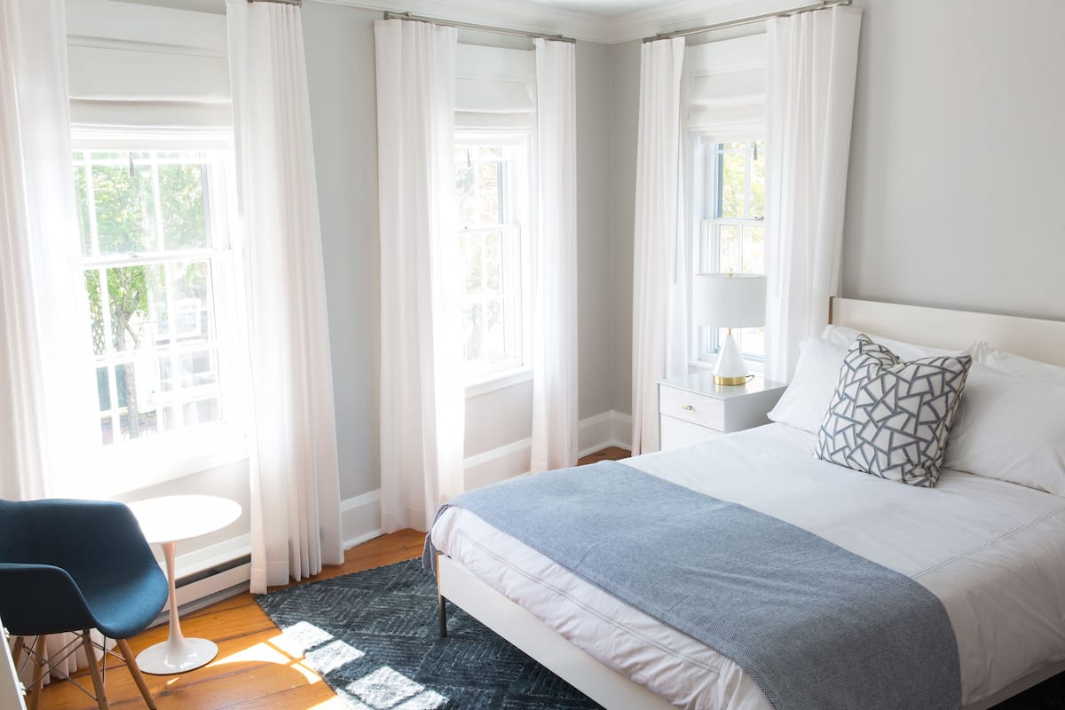 Ein Schlafzimmer mit Bett, Nachttisch, Lampe, Stuhl und einem Fenster mit Vorhängen.