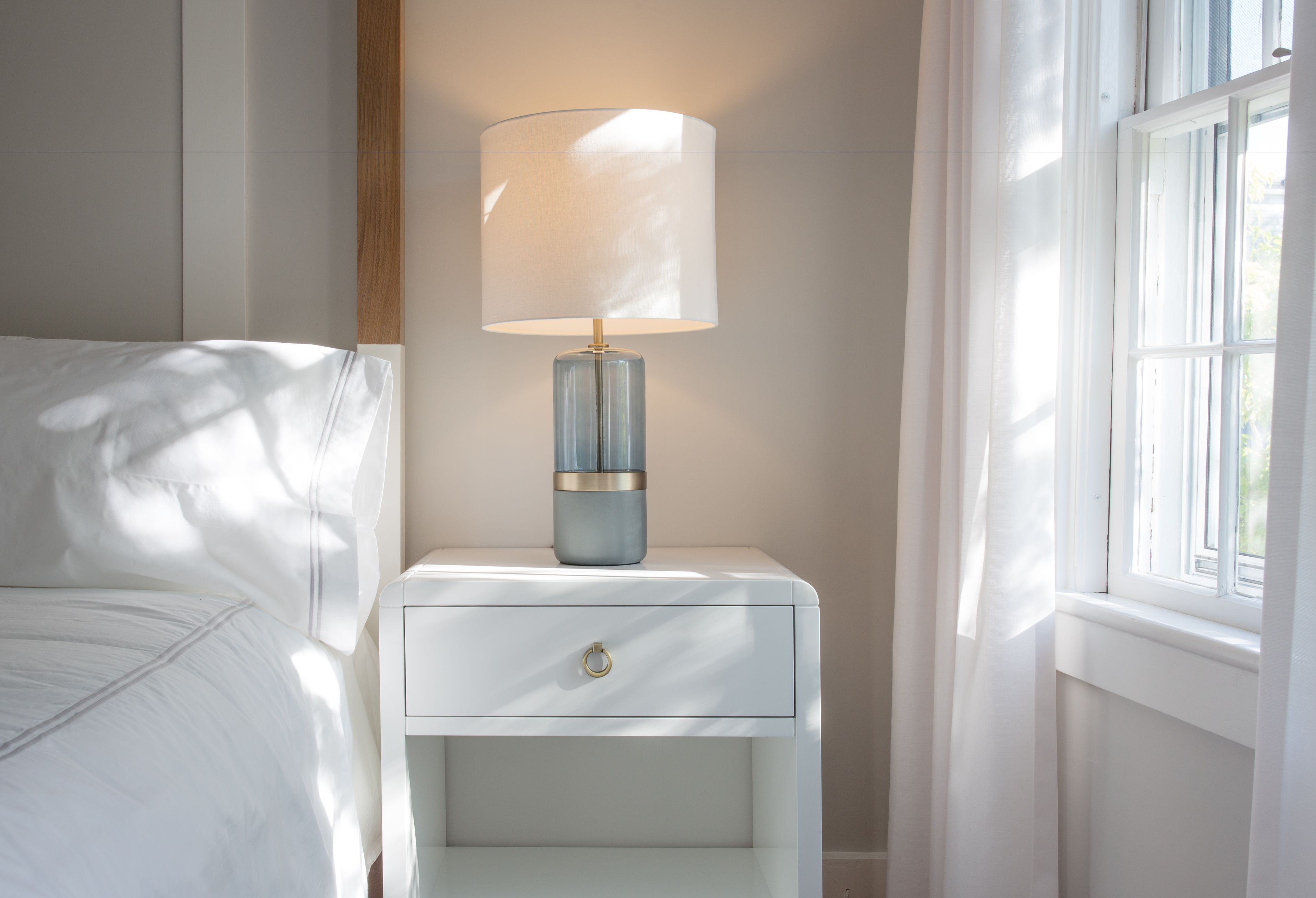 A white bedside table with a lamp, a drawer, and a window with curtains.