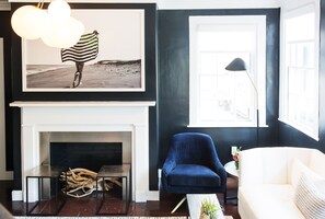 Una sala de estar con chimenea, un sillón azul, un sofá blanco y un cuadro grande enmarcados con una escena de playa.