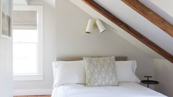 Quarto com teto inclinado, cama com roupa de cama branca, almofada estampada e uma pequena mesa redonda.