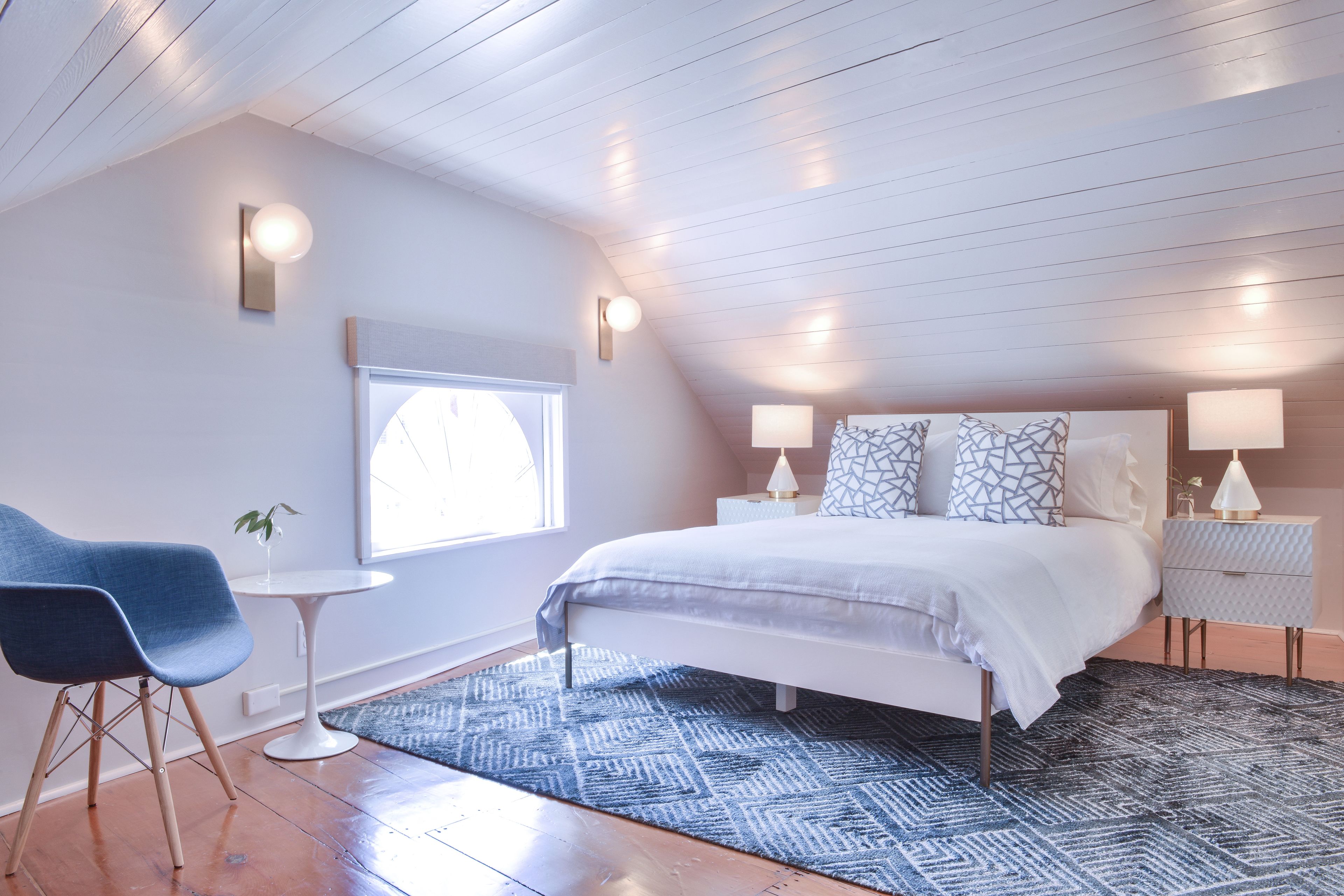 A modern bedroom with a bed, bedside tables, a chair, a small table, and a window.
