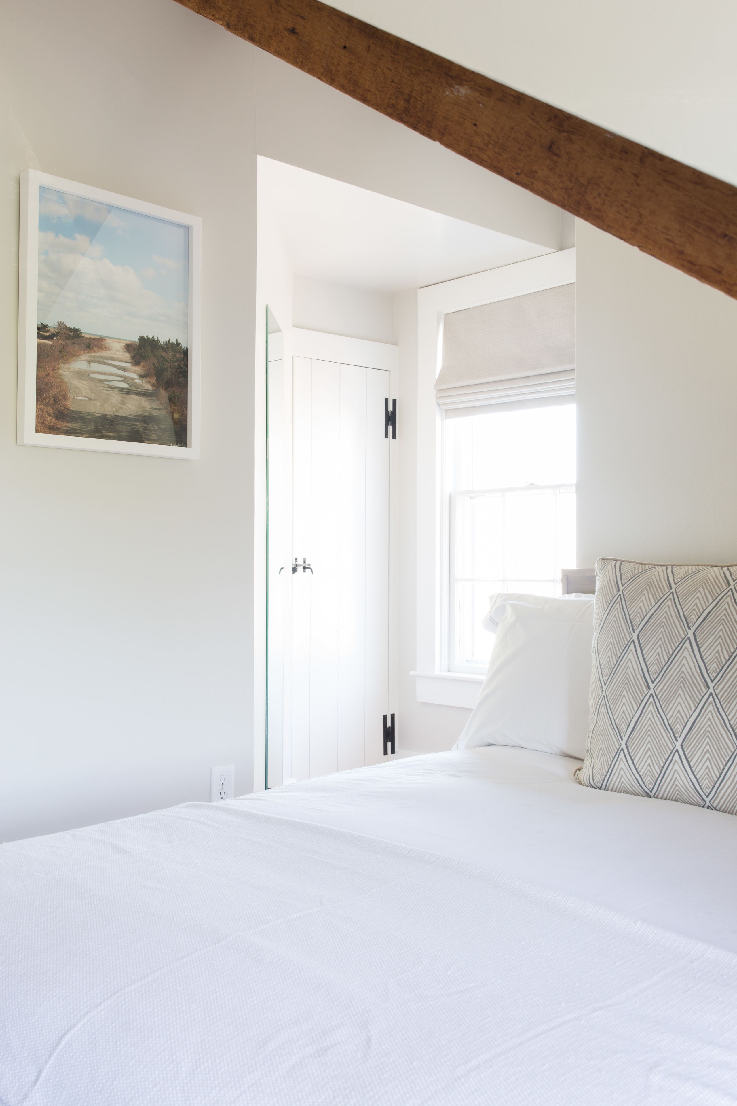 A bedroom with a white bed, a patterned pillow, a painting on the wall, and a window with blinds.