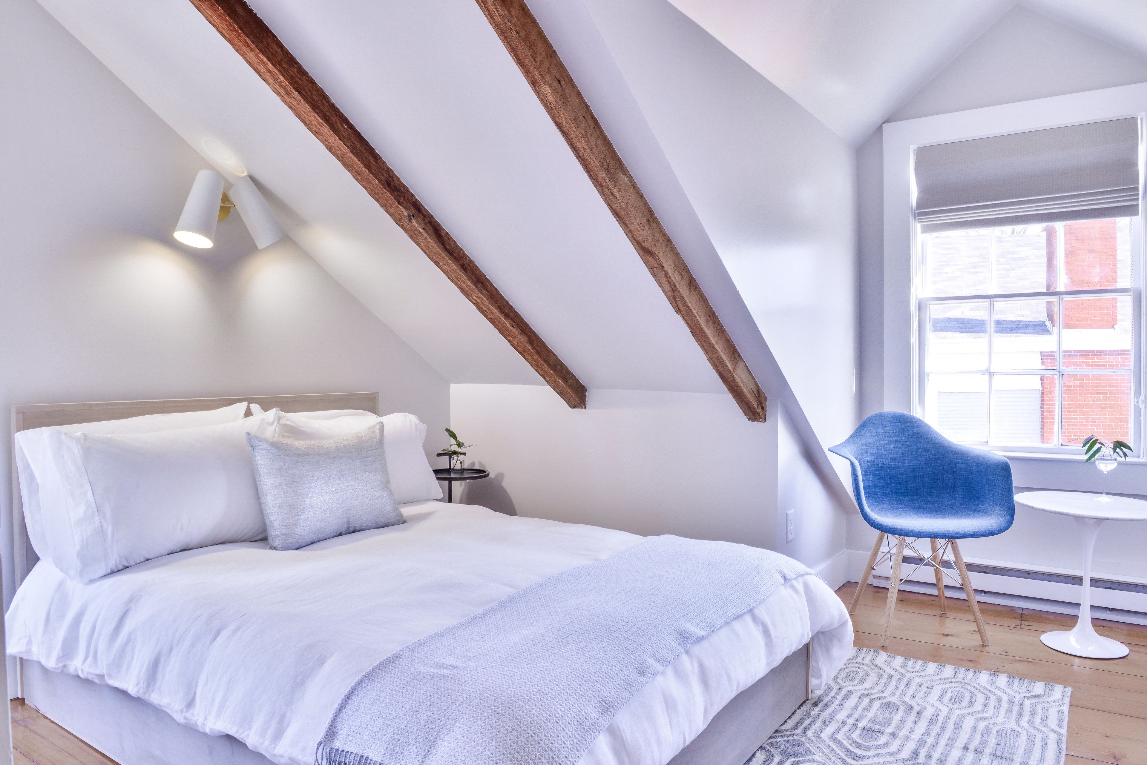 Ein Schlafzimmer mit schrägem Dach, einem Bett mit weißer Bettwäsche, einem blauen Sessel und einem kleinen Tisch.