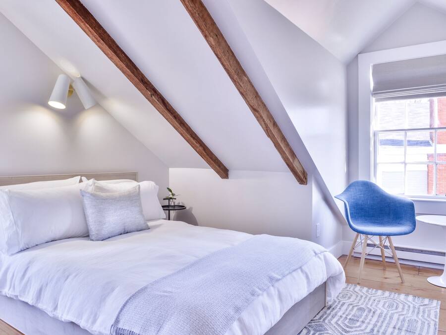 Ein Schlafzimmer mit schrägem Dach, einem Bett mit weißer Bettwäsche, einem blauen Sessel und einem kleinen Tisch.