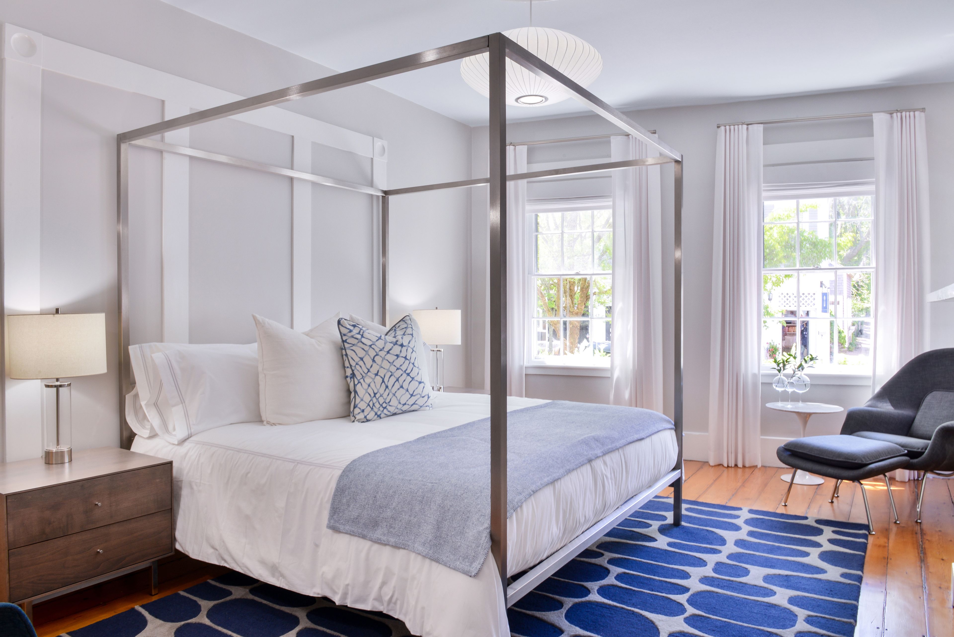 Un lit à baldaquin avec une couvre-lit et des oreillers blancs, une table de chevet avec une lampe, un tapis à motifs bleus et une vue sur l’extérieur par une fenêtre.