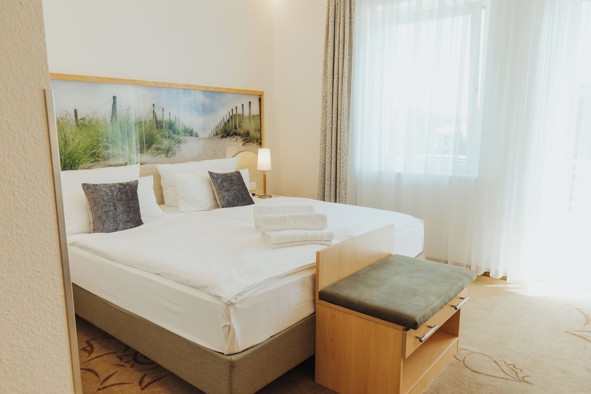 A neatly made bed with white linens and pillows, a bedside table with a lamp, and a painting of a beach scene above the bed.