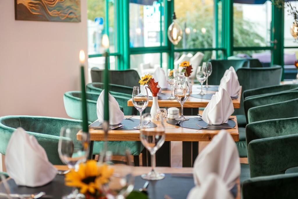 A restaurant with green upholstered chairs, wooden tables, and a view through large windows.