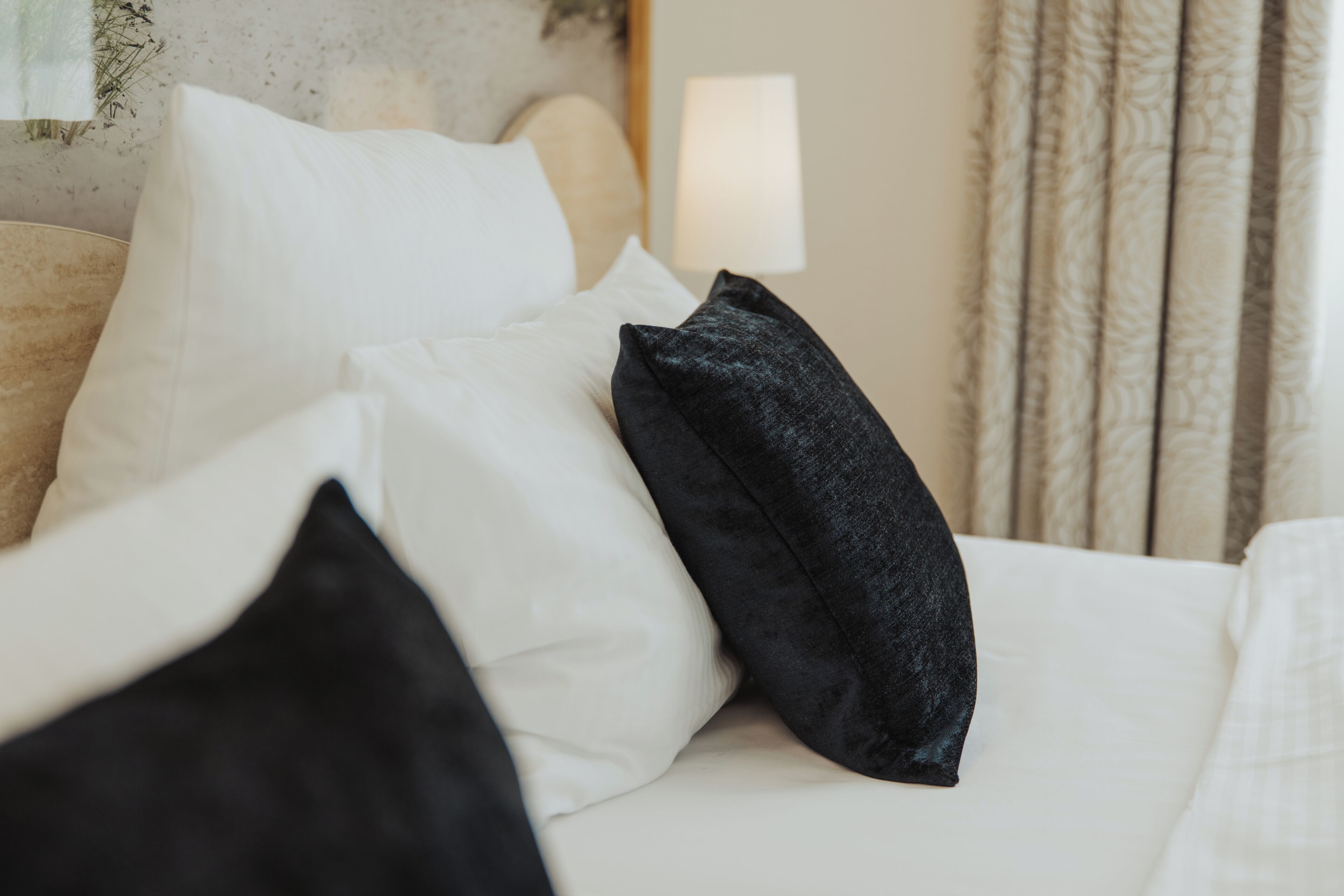 Ein ordentlich bezogenes Bett mit weißen und schwarzen Kissen, eine Lampe daneben und ein gemusterter Vorhang im Hintergrund.